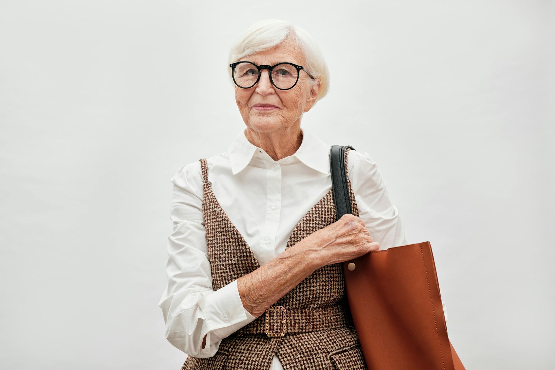 Woman with glasses and a brown tote bag, wearing a white shirt and patterned vest, stands against a white background.