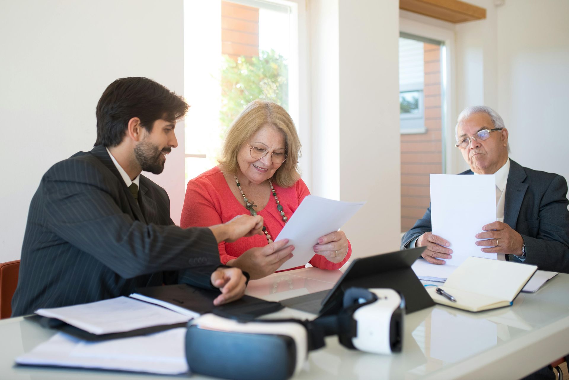 Three people review documents at a table. A man points at a paper as a woman smiles. Another man holds paper, looking on.