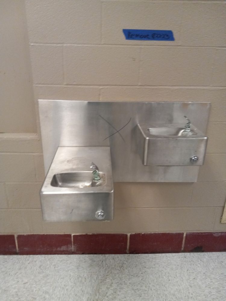 Wall-mounted stainless steel drinking fountains on a tiled restroom wall, one low and one higher for accessibility