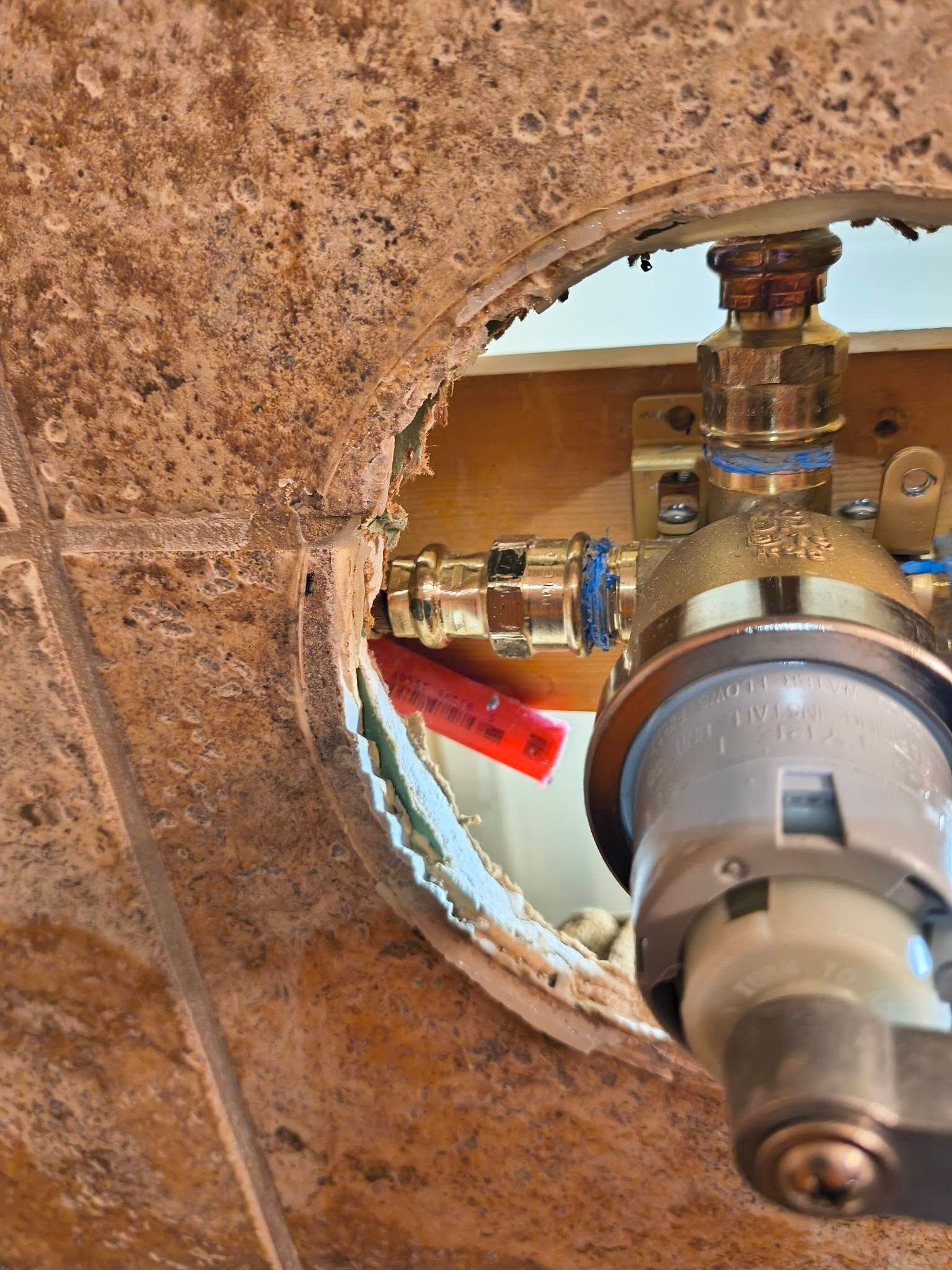 Close-up of a rusty wall opening revealing a pipe valve and copper plumbing inside.