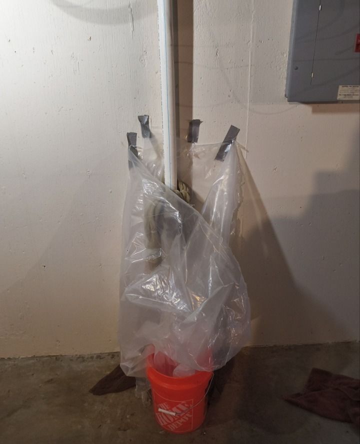 A clear plastic bag hangs on a white pipe above a red bucket in a basement corner.