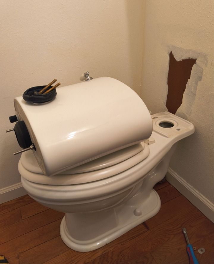 Broken white toilet with a lifted tank lid and exposed wall damage in a bathroom corner