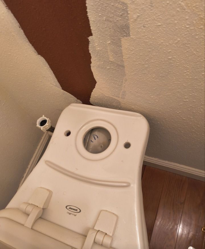 White toilet tank on floor beside damaged wall with exposed brown patch and loose plumbing pipe.