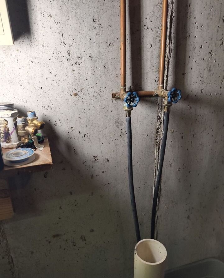 Plumbing pipes with blue valves on a concrete wall above a white bucket in a dim utility room