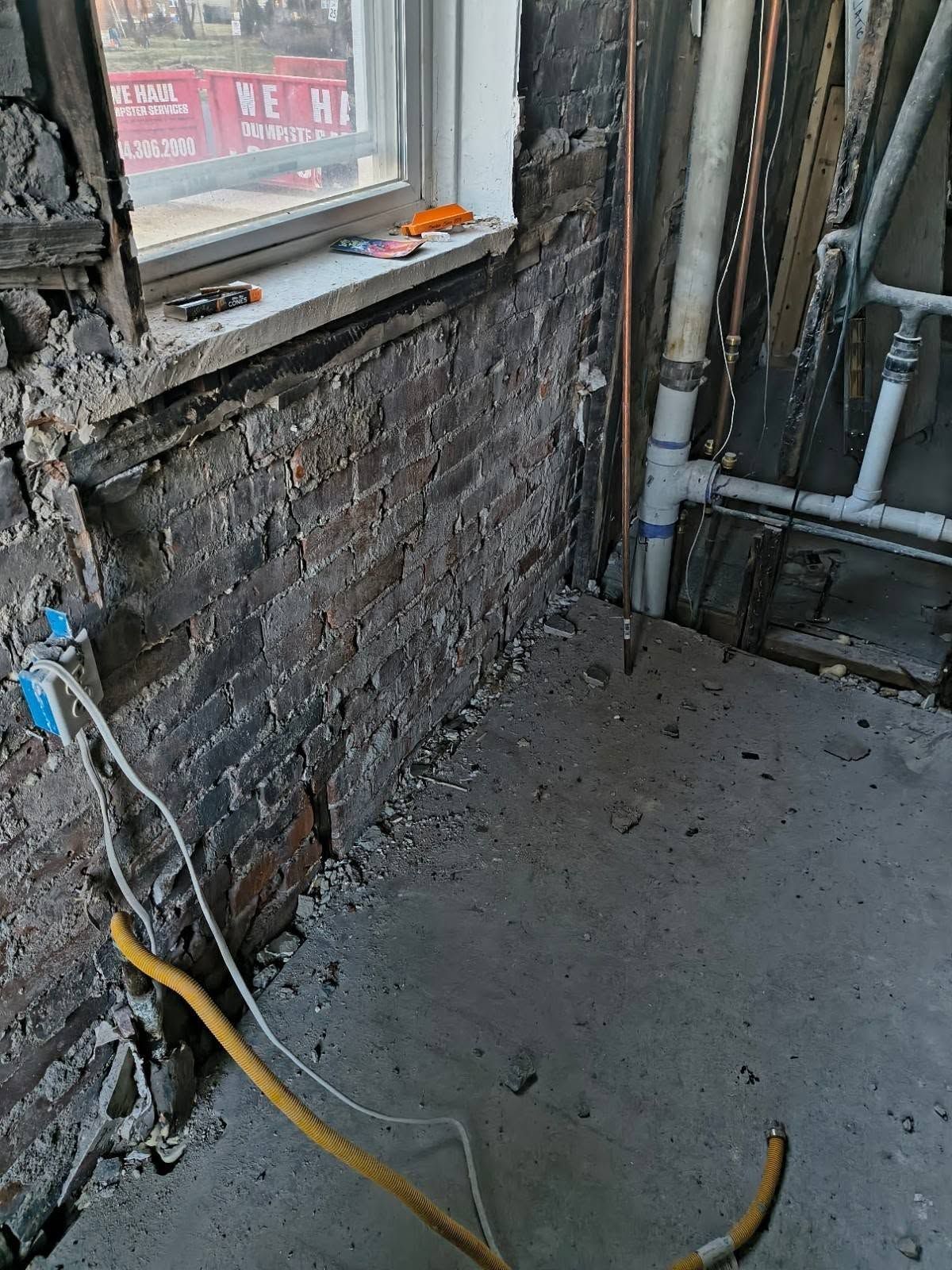 Partially renovated room with exposed brick wall, window, and exposed pipes on a concrete floor.