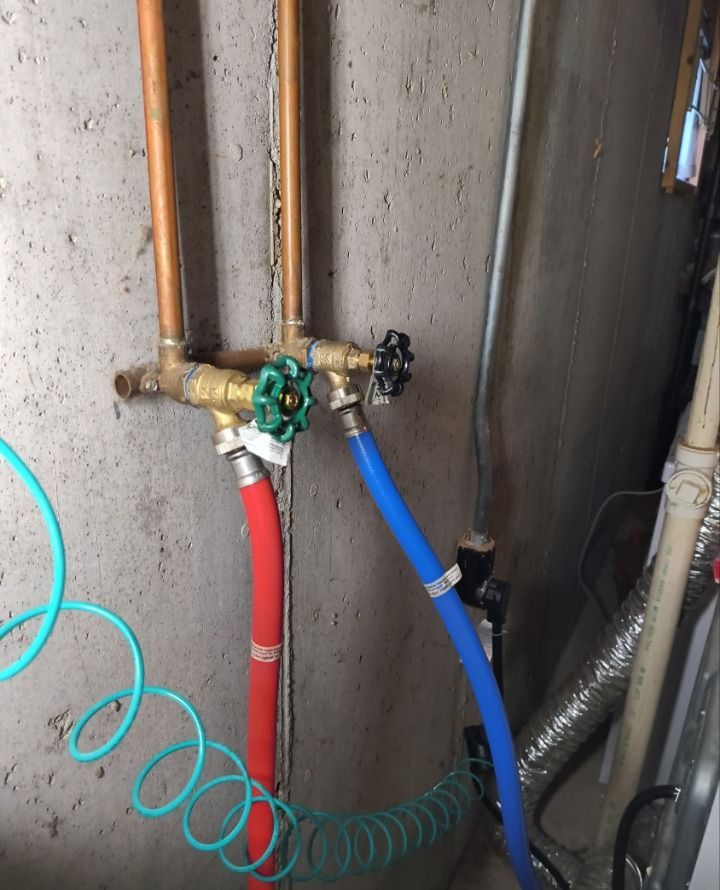 Plumbing manifold with red and blue hoses and green valve handles on a concrete wall