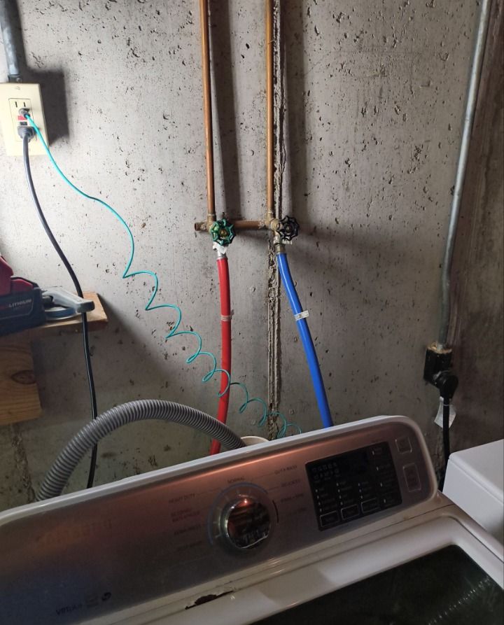 Laundry area with washer, exposed pipes, and blue hose against a concrete wall