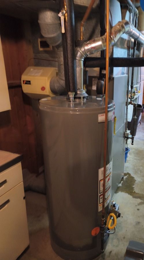 Tall silver water heater with pipes and controls in a utility room, with a small water spill on the floor.