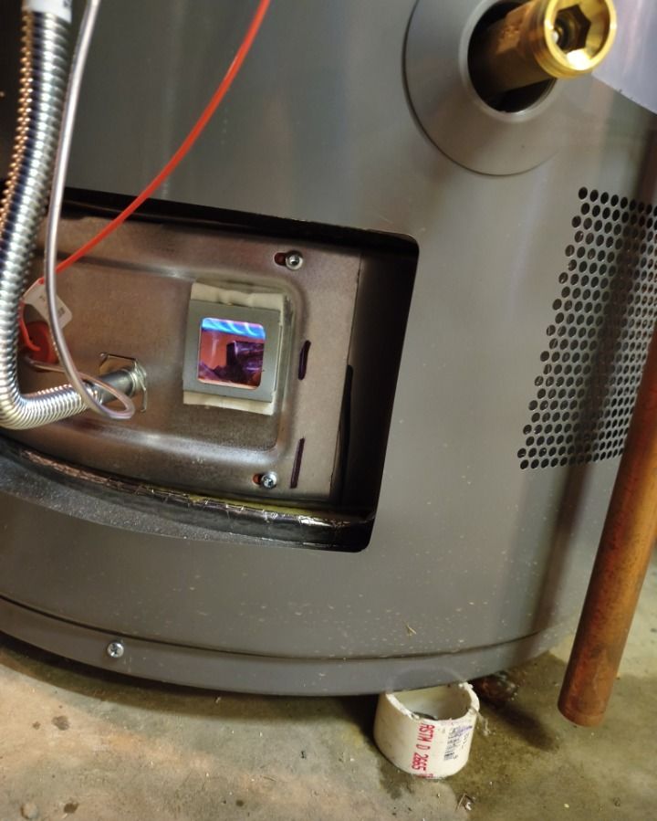 Close-up of a metal appliance with a small viewing window and a pipe, with a cup on the floor nearby.
