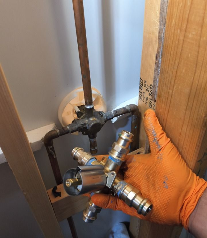 Gloved hand holding a metal plumbing valve assembly beside exposed pipes and a wall opening