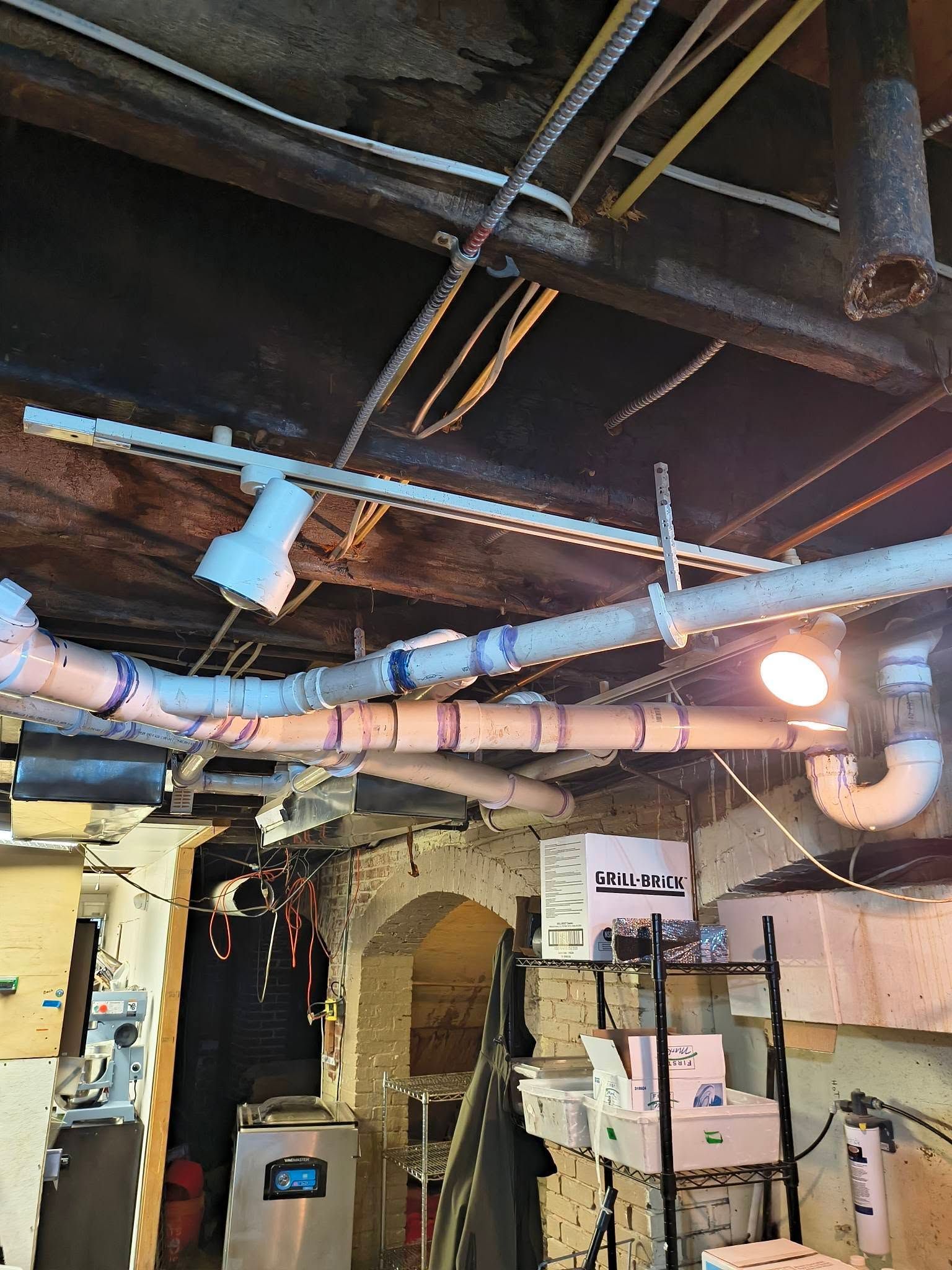 Basement utility room with exposed pipes, wires, and a bright hanging light over stacked equipment.