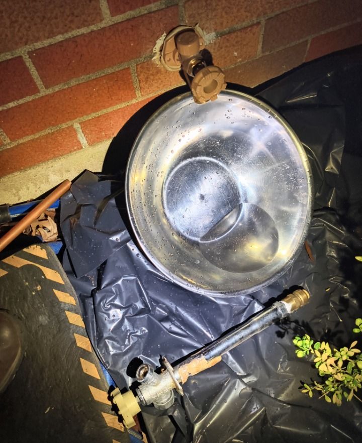 Large metal bowl or sink propped against a brick wall, with black bags and pipes around it.