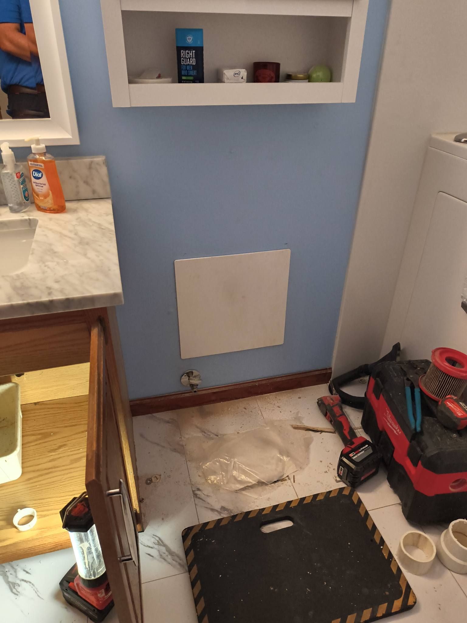 Bathroom wall with a cut-out panel, tools, and debris on the floor during repair.