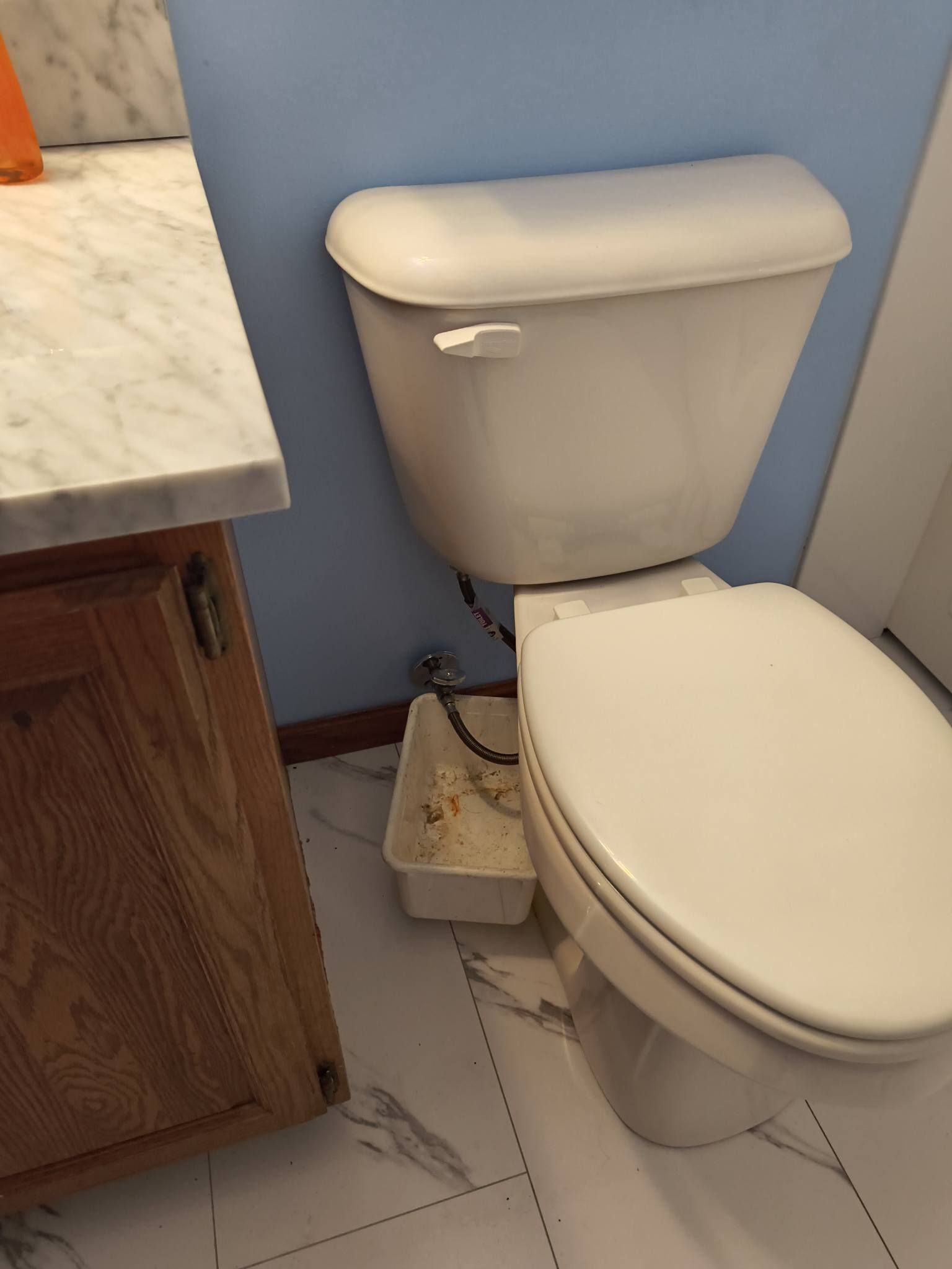 Small bathroom with toilet, beige cabinet, white tile floor, and a utility bin beside the toilet.