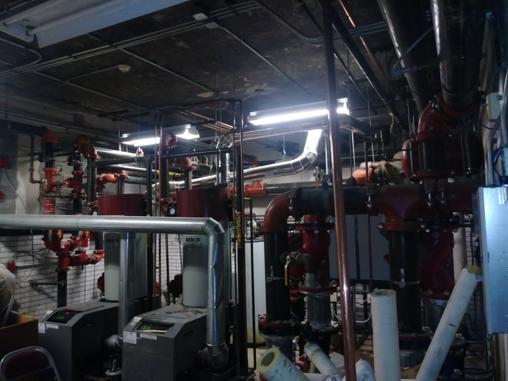 Dim industrial basement with red pipes, ducts, and machinery under fluorescent lights