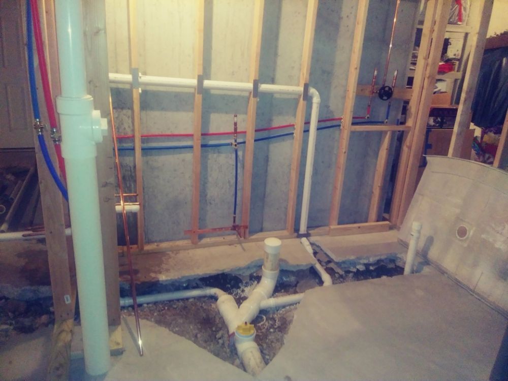 Exposed wall framing and plumbing in a bathroom under renovation, with a concrete floor and exposed pipe drain