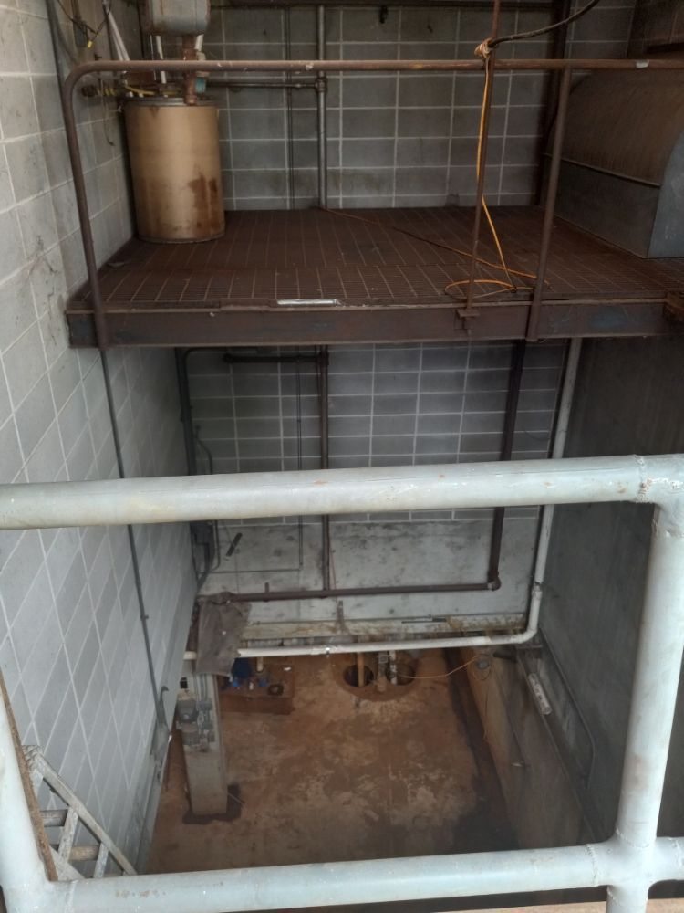 Industrial shaft with metal catwalk, pipes, and a rusty lower pit behind safety rails