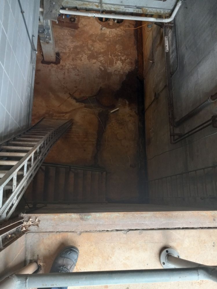 Looking down a narrow concrete shaft with a ladder and rusty rock wall at the bottom