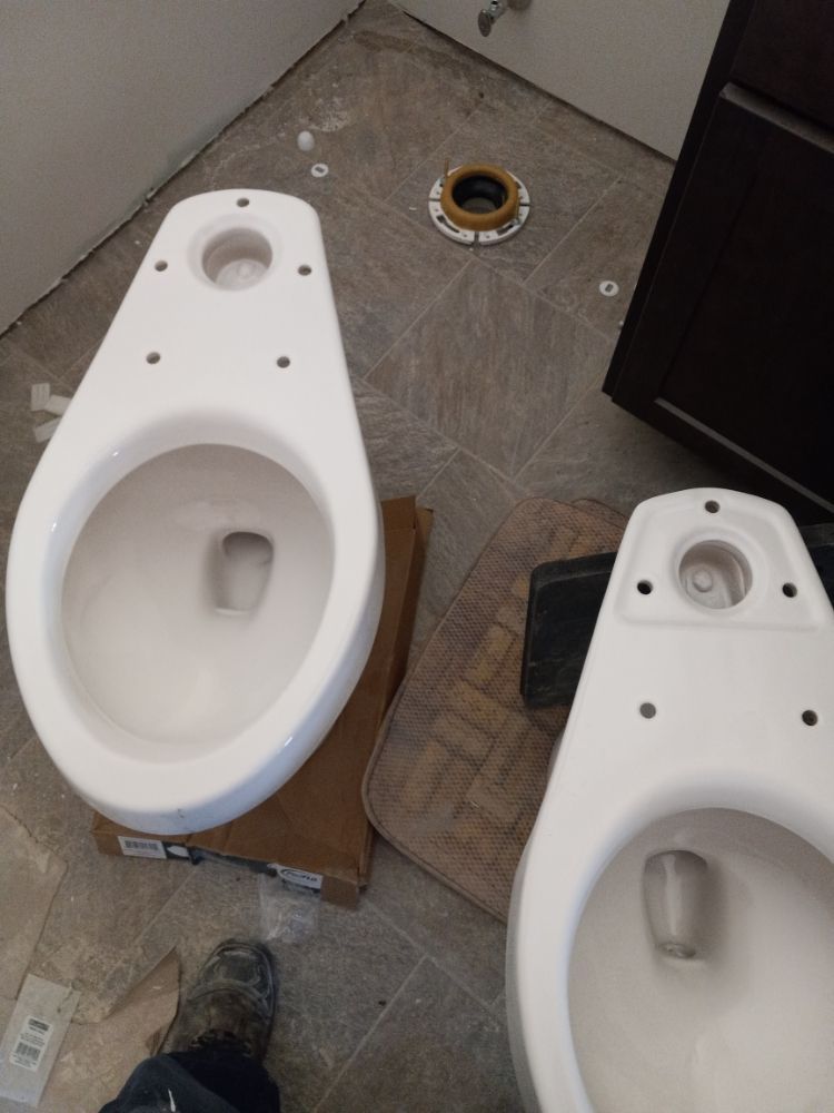 Two white toilet bowls on a floor, with a small circular drain ring nearby in a bathroom workspace.