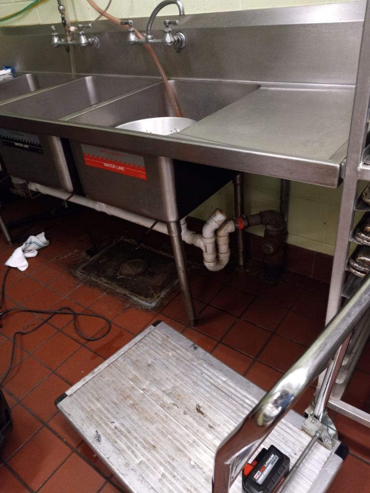 Commercial kitchen sink with stainless steel counter and tiled floor, plus a floor mat and cleaning cord underneath