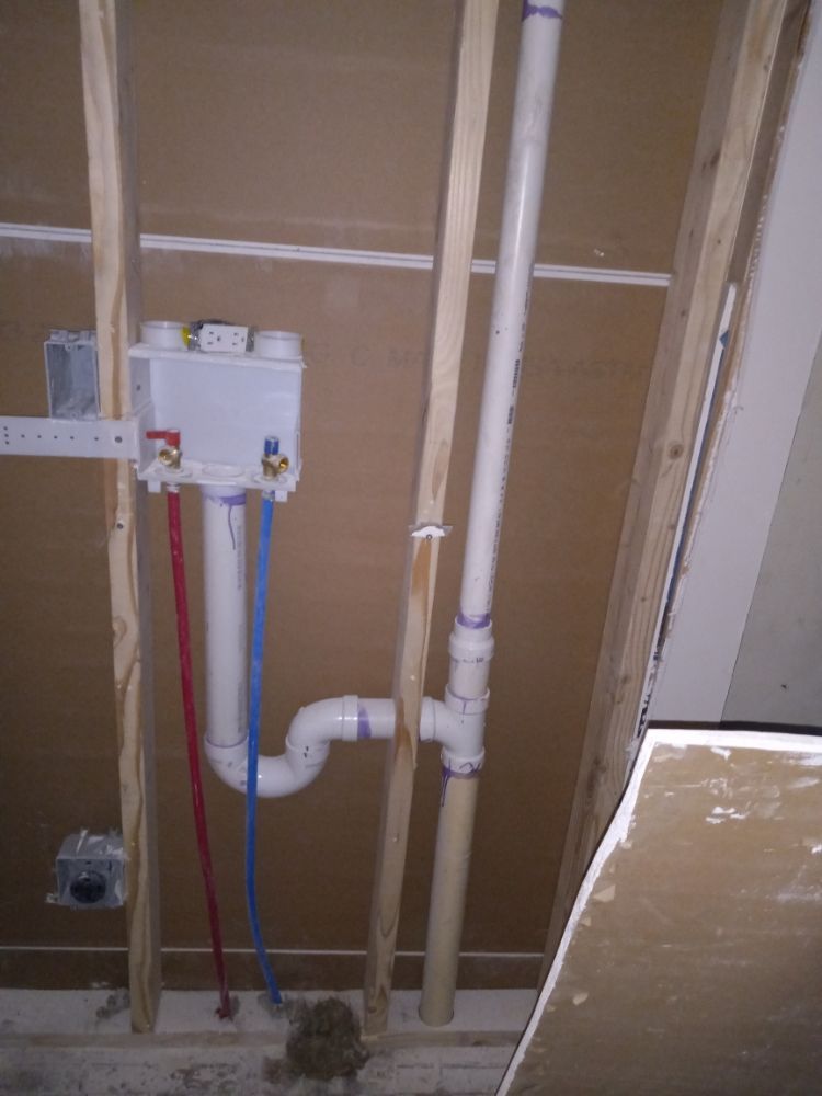 Open wall with exposed plumbing, white pipes, and a utility box in a construction area.