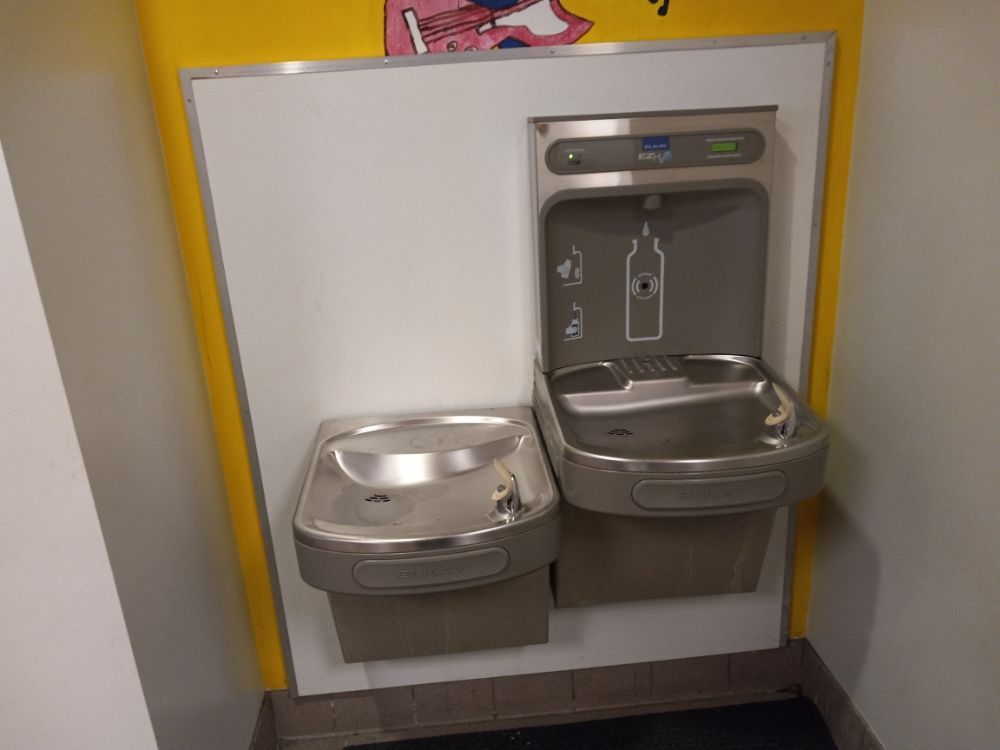 Two stainless steel drinking fountains mounted on a yellow wall, one low and one standard height.