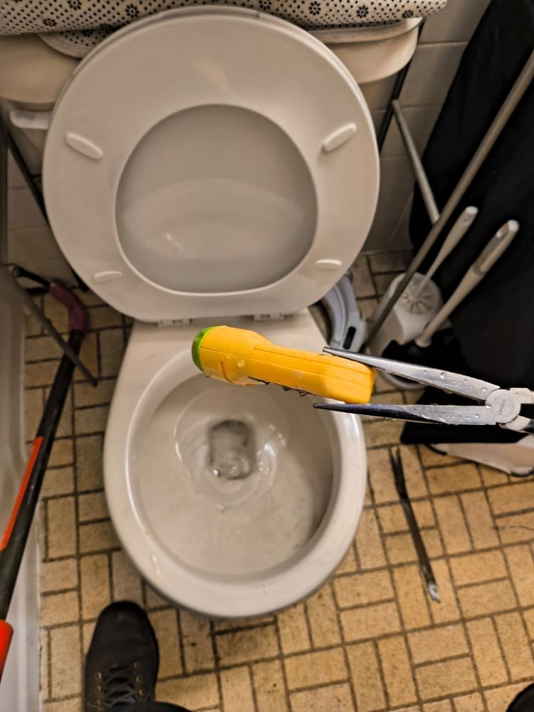 Open toilet seat with a yellow-handled brush cleaning the bowl in a small tiled bathroom
