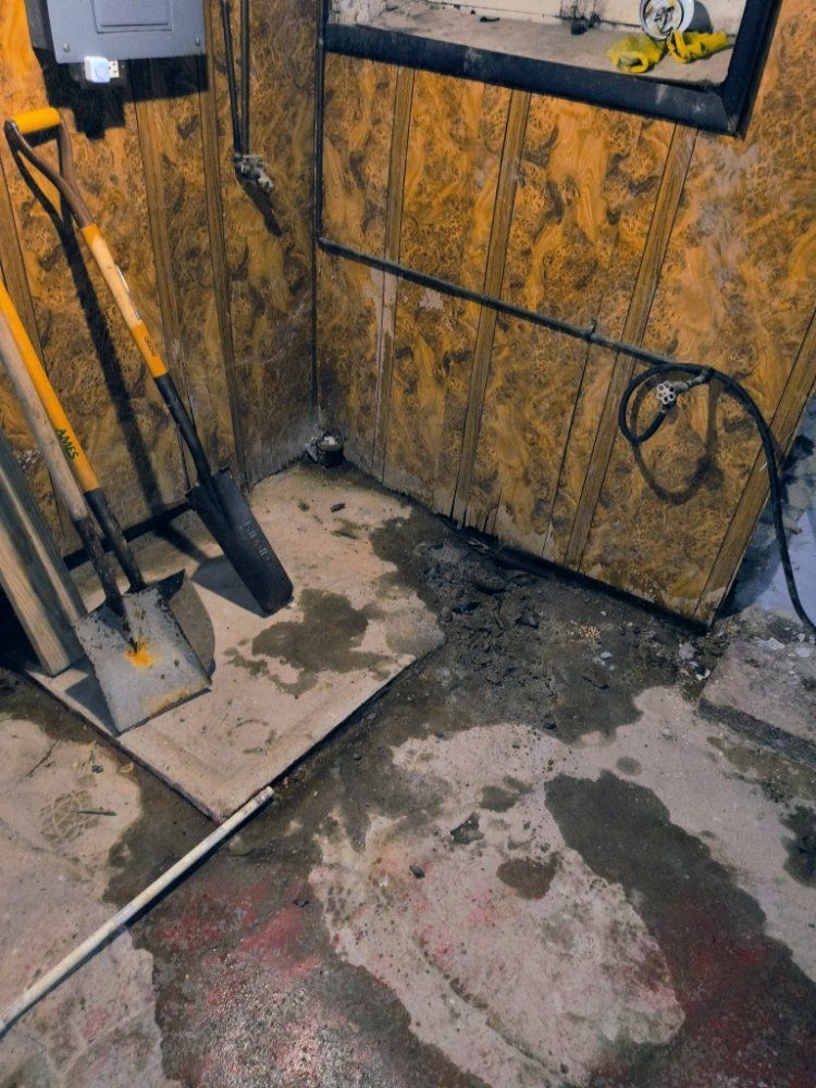 Shovel and tools leaning against a wall in a dusty, cluttered utility room with stains on the floor.
