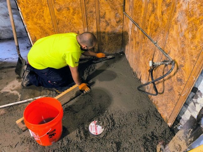 Worker troweling wet concrete beside an orange bucket in a plywood-walled corner