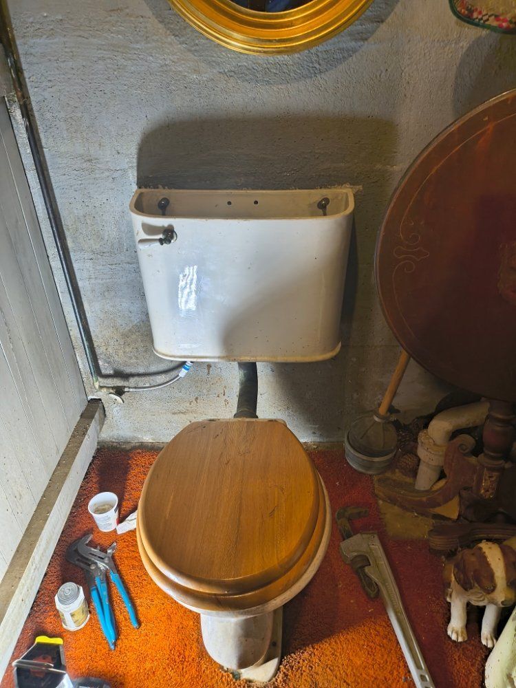 Small bathroom with a toilet, wall-mounted tank, orange bath mat, and cleaning tools on the floor.