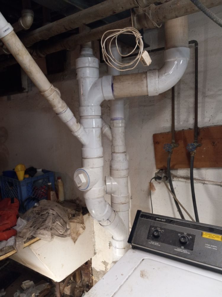 White PVC drainage pipes and hose connections in a basement laundry area near a washer/dryer.