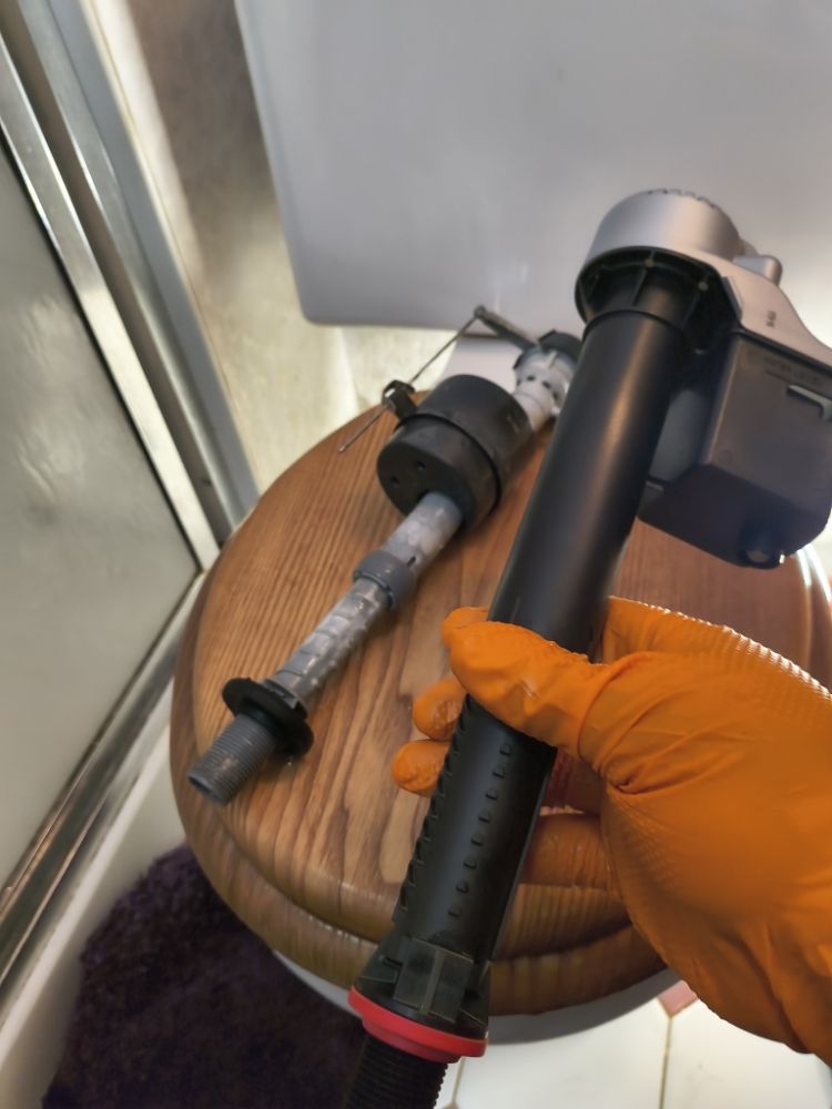 Orange-gloved hand holding a black power tool with attachments on a wooden stool by a window