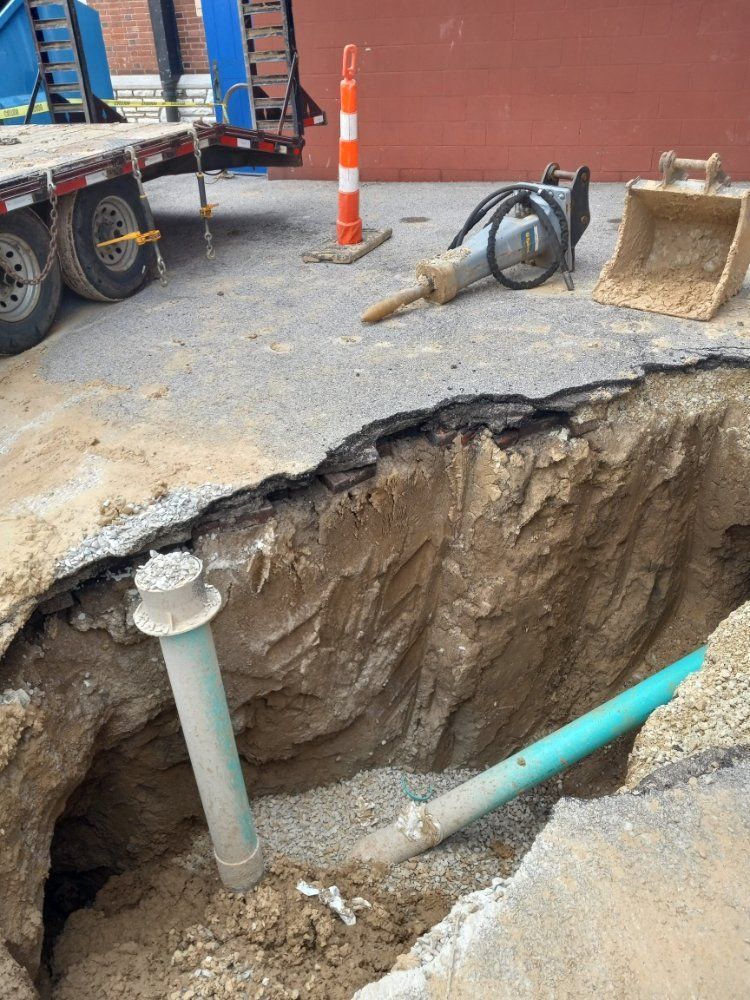 Collapsed road trench with exposed pipes, construction truck, traffic cone, and bucket on a street worksite