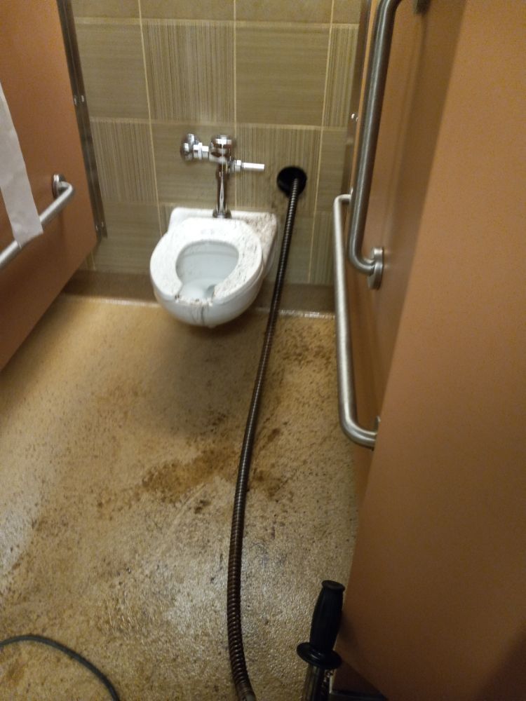 Public restroom with a toilet, sink spray hose, and wet stained floor beside a shower area