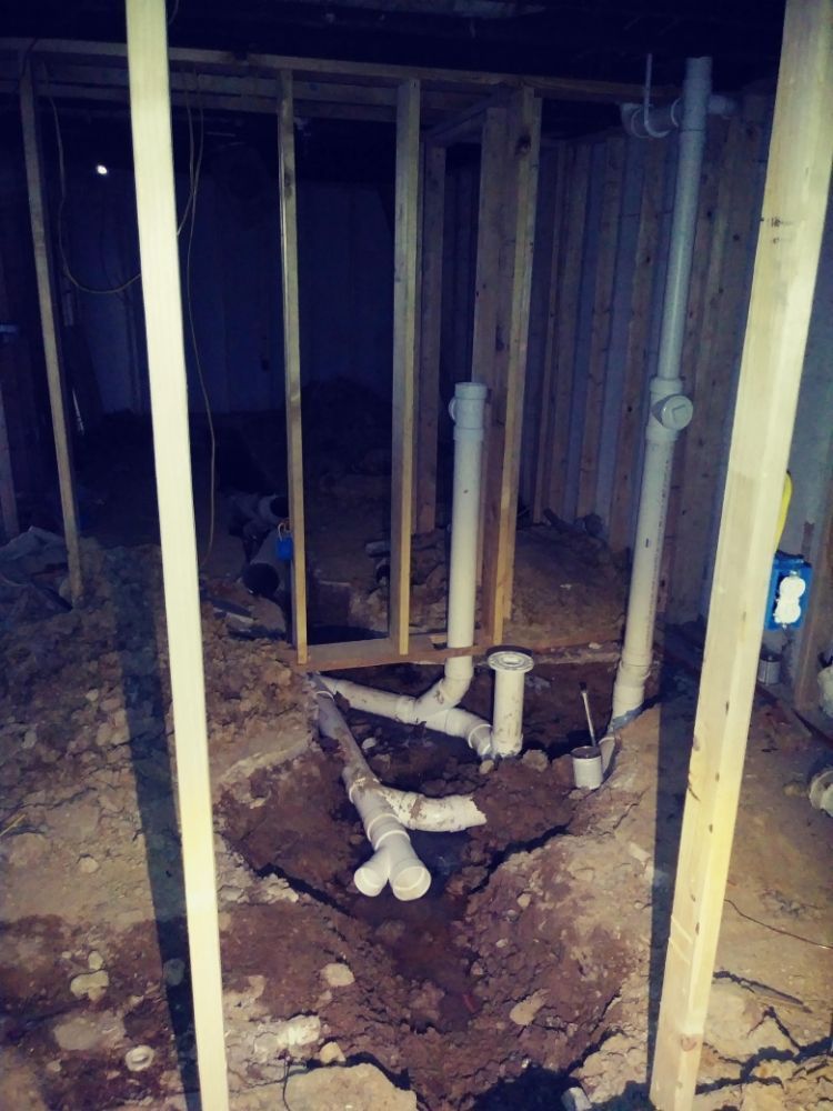 Unfinished basement with exposed wooden studs and rough plumbing pipes in a dirt floor trench.