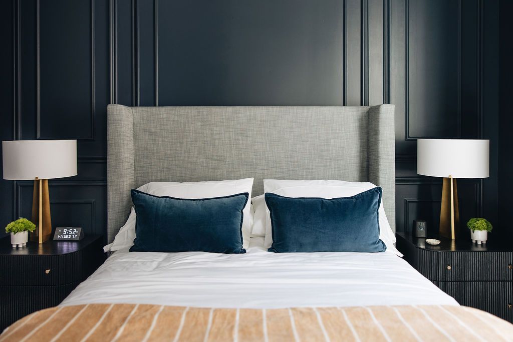 Bedroom with a gray upholstered headboard, blue pillows, and dark blue paneled walls; two nightstands flank the bed with gold-based lamps. Purple Martin Hotel