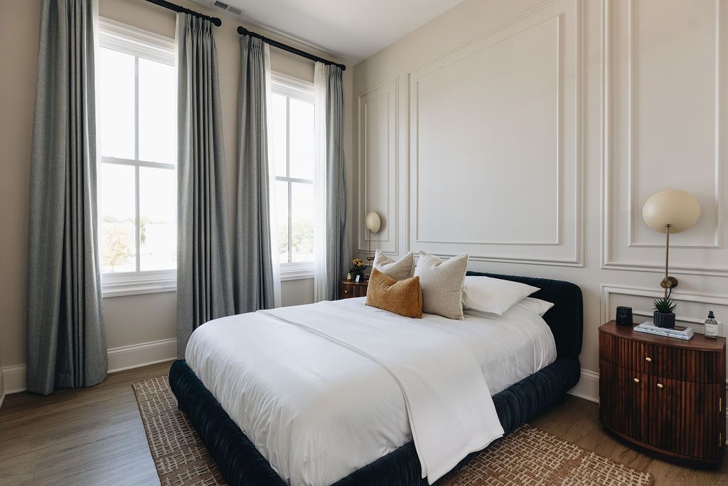 Bedroom with white walls, two windows with gray curtains, bed with white bedding, and wood nightstand. Purple Martin Hotel