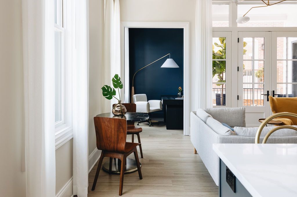 Living room interior with hardwood floors, white walls, and a view of a dark blue office.