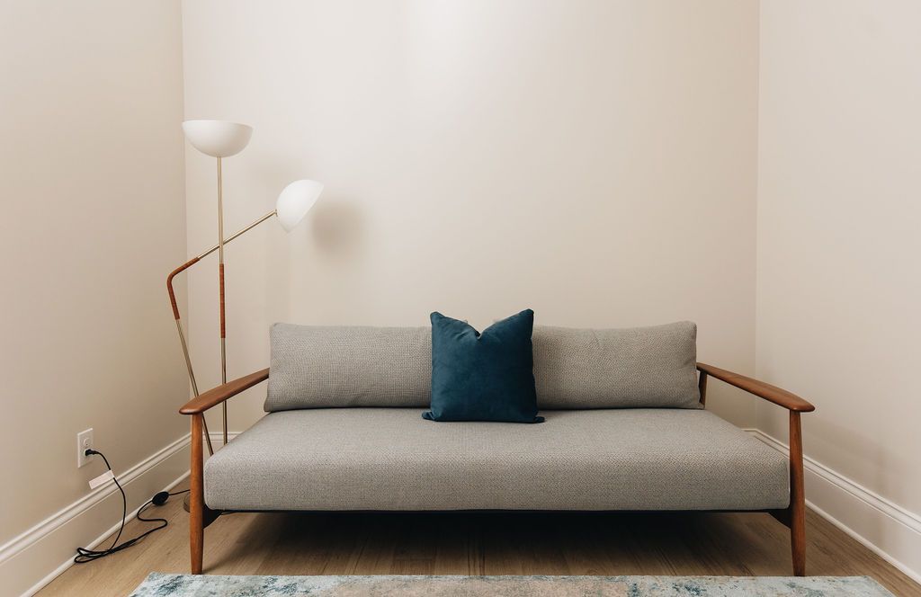 Sofa with blue pillow and wooden frame, floor lamp, and cream walls. Purple Martin Hotel