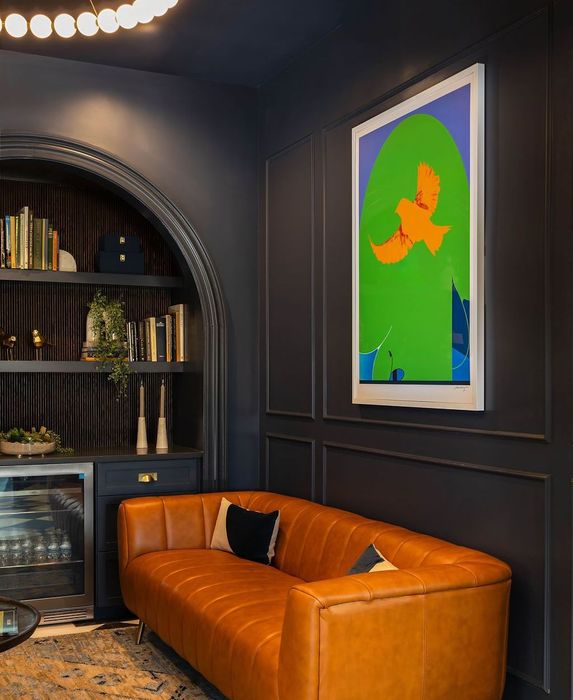Cozy room with orange leather sofa, a vibrant artwork, and a built-in bookshelf against dark blue walls. Purple Martin Hotel