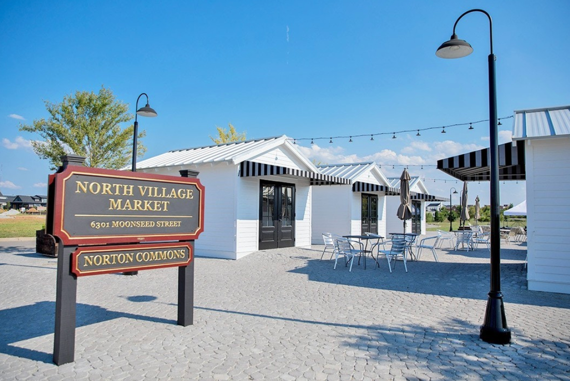 North Village Market sign in front of white buildings with black awnings, outdoor tables and lights, blue sky.