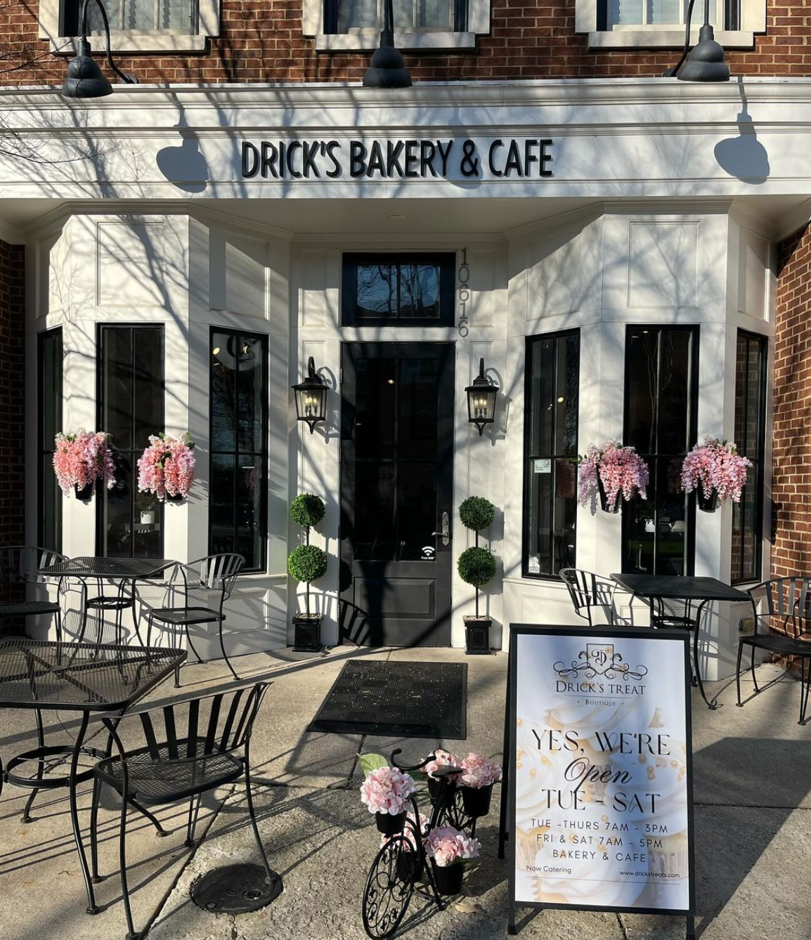 Exterior of Drick's Bakery & Cafe with outdoor seating and flower displays. Sunlight casts shadows. Sign reads:
