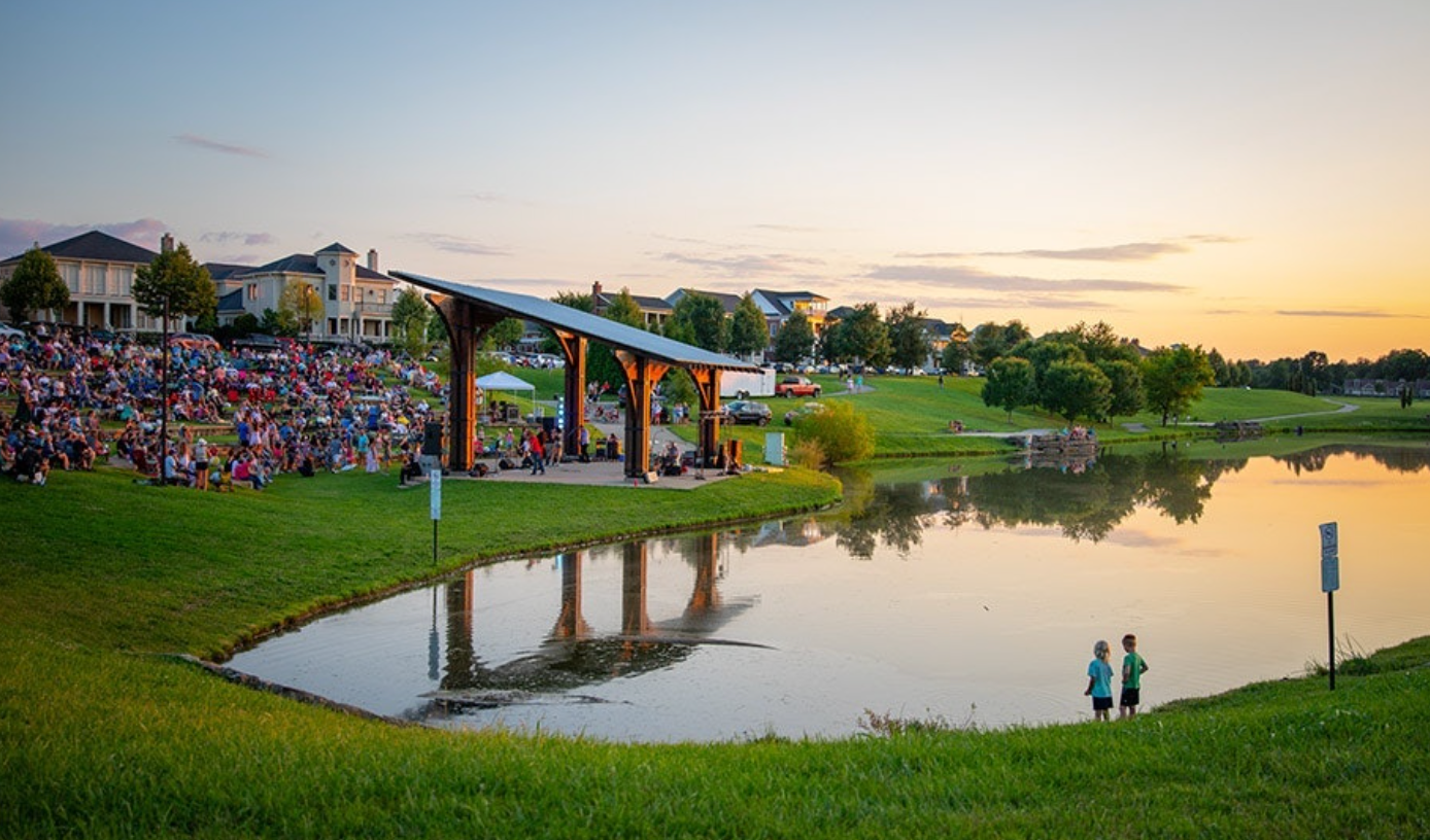 Lakeside park at sunset with a pavilion, a crowd seated on the lawn, and two people standing near the water. Purple Martin Hotel