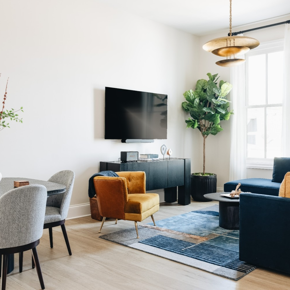 Living room with a TV, blue sofa, yellow chair, round table, and a large plant. Purple Martin Hotel