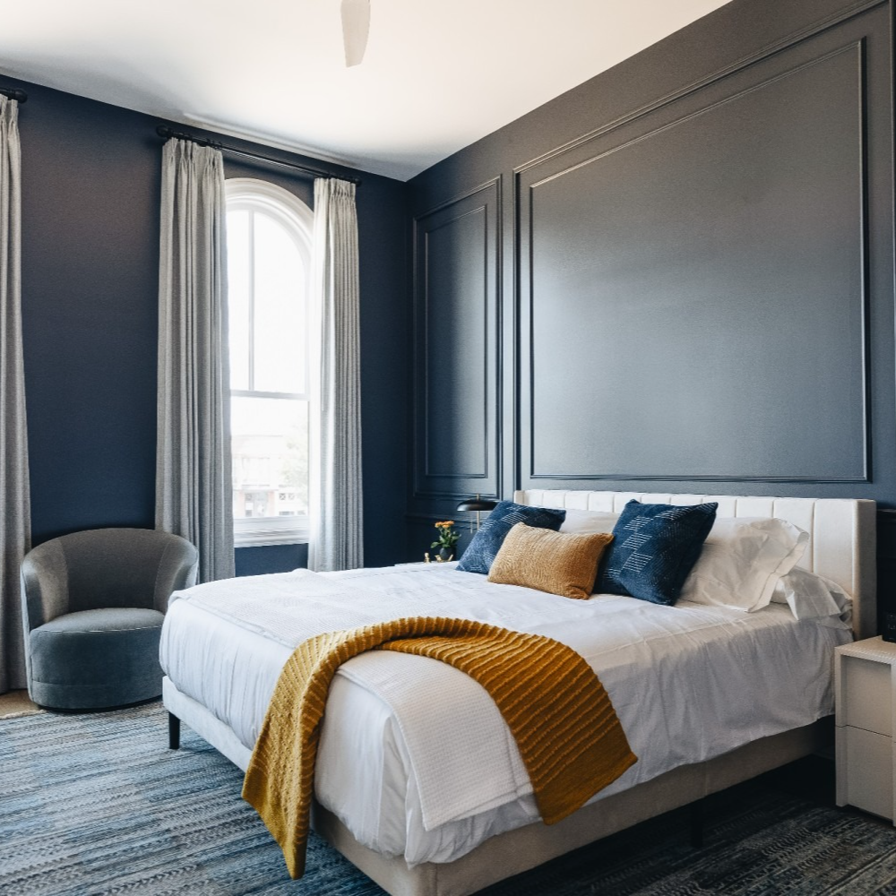 Dark blue bedroom with white bed, gray armchair, and golden blanket. Purple Martin Hotel