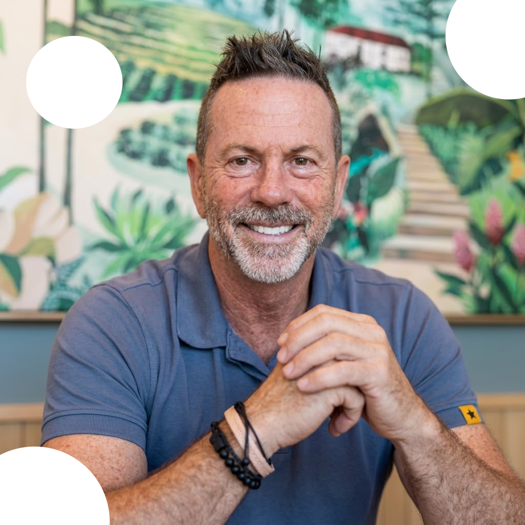 Smiling man in a blue polo shirt sitting with hands clasped against a leafy mural background