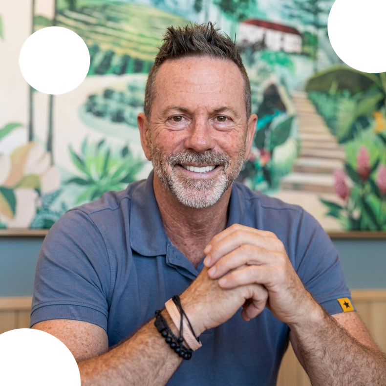 Smiling man in a blue polo shirt sitting with hands clasped against a leafy mural background