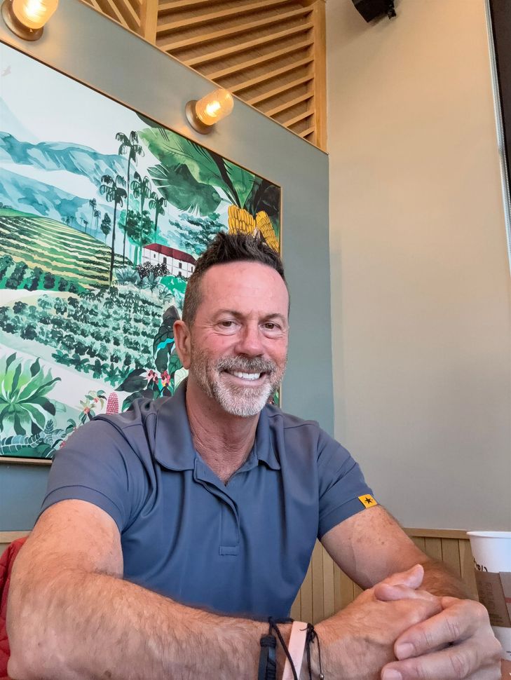 Smiling man in a blue polo shirt sitting with hands clasped against a leafy mural background