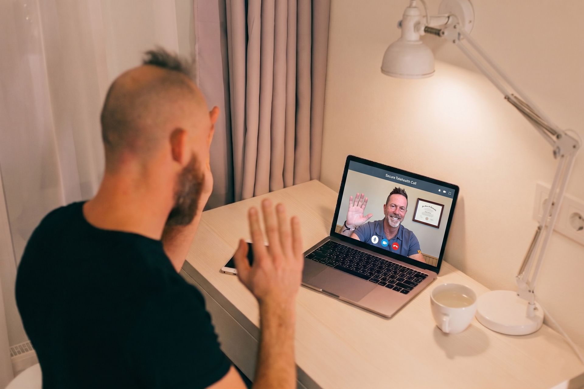 Person on a video call at a desk with a laptop, lamp, and coffee mug in a small home office.