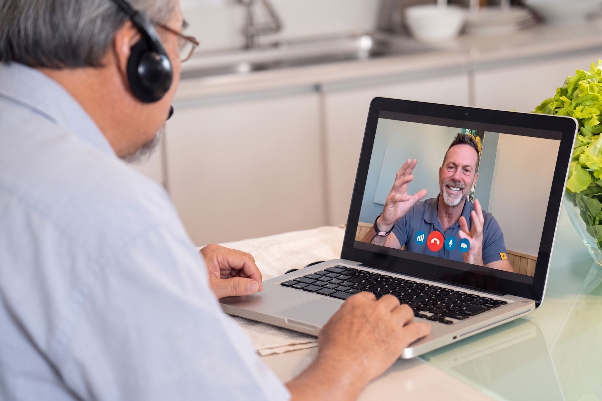 Person wearing headphones on a laptop video call with a man gesturing from a kitchen table